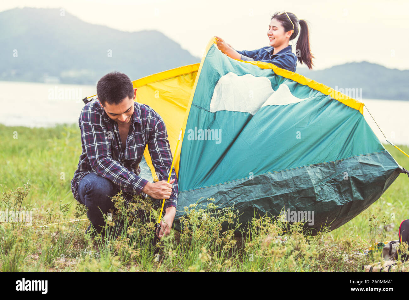 Two Asian couples prepare pitch camping tent to stay outdoors meadow overnight for honeymoon camping picnic. People lifestyle and Valentine day love c Stock Photo