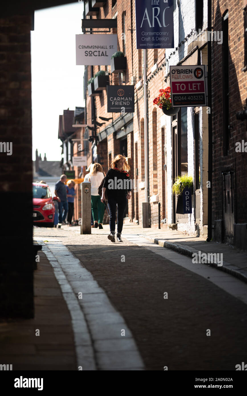 A woman (age 30s) walks towards purposefully towards Leen Lane, a small lane overshadowed by buildings in central Chester. An open sunlit area behind. Stock Photo