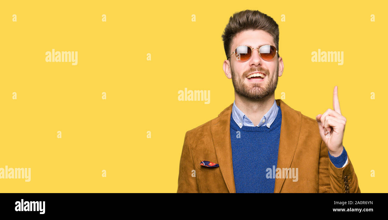 Young handsome elegant man wearing sunglasses pointing finger up with ...