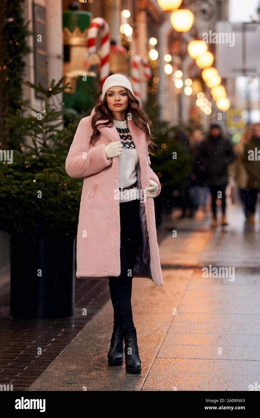 Outdoors lifestyle fashion portrait of beautiful brunette woman walking christmas decorated city street. Smiling woman. Female wearing pink stylish fu Stock Photo