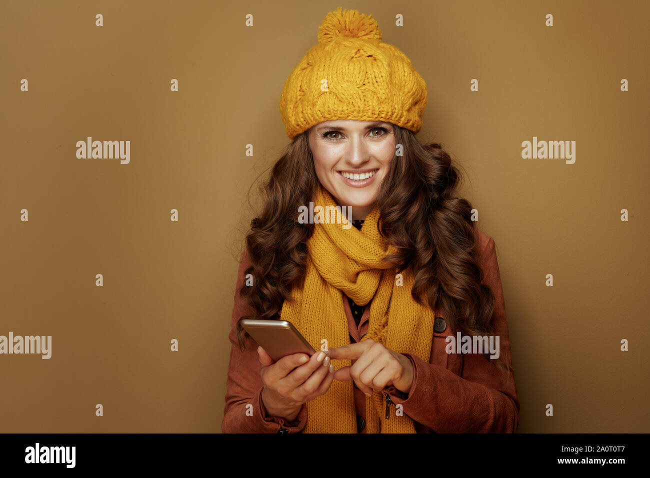 smiling trendy woman in yellow beret and scarf with smartphone hanging in social media and against bronze background. Stock Photo