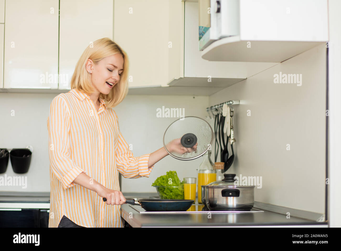 Happy beautiful caucasian blonde woman standing in kitchen near stove with spatula, cooking eggs in frying pan in morning. Female student cooking brea Stock Photo
