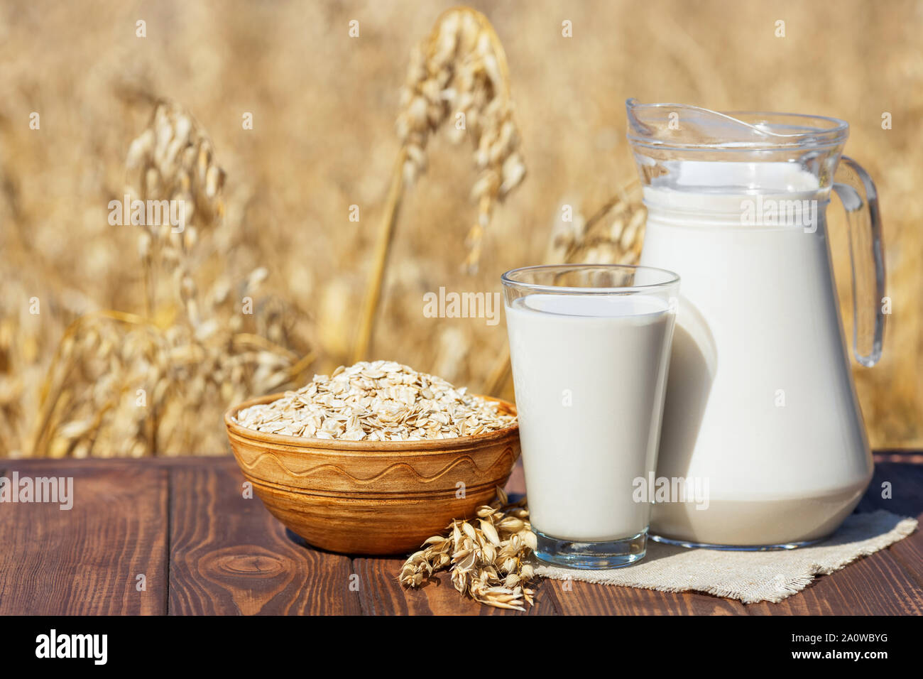 vegan oat milk in glass and jug with uncooked oatmeal in bowl on table over against ripe cereal field Stock Photo