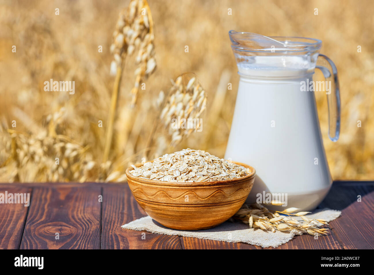 vegan oat milk in glass jug with uncooked oatmeal in bowl on table over against ripe cereal field Stock Photo