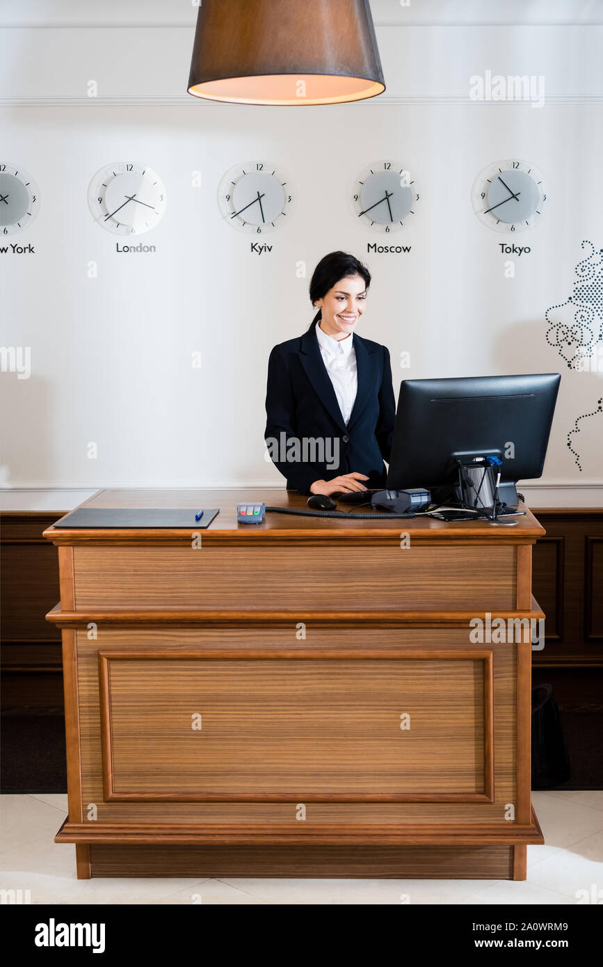 cheerful brunette receptionist in formal wear looking at computer ...