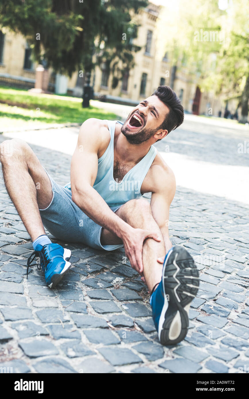sportsman suffering from pain and screaming while sitting on pavement ...