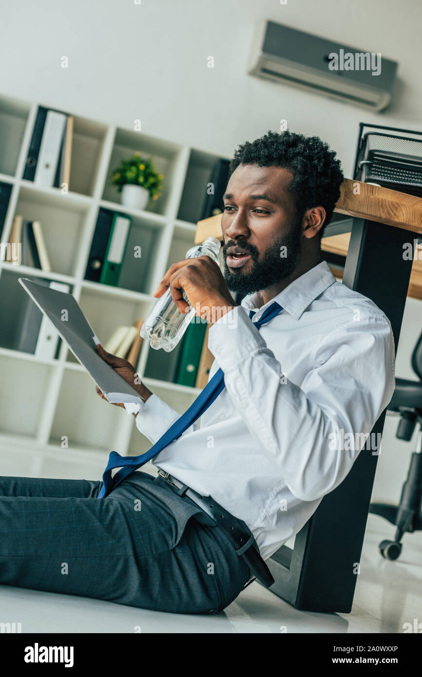 young african american businessman sitting on floor and drinking water  while suffering from heat in office Stock Photo - Alamy, image size:867x1390