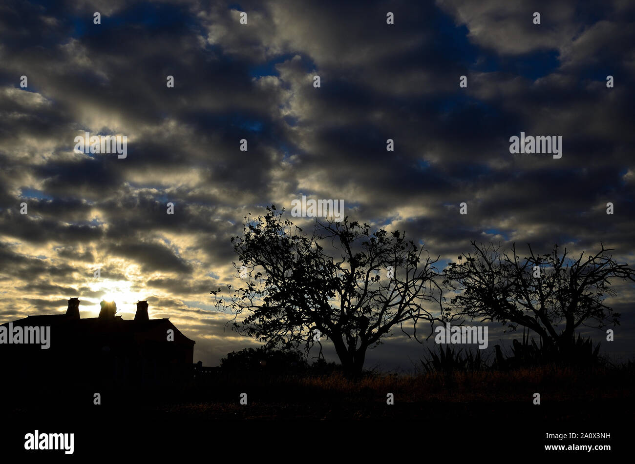 Backlit countryside landscape at sunrise with cloudy sky, house and trees Stock Photo