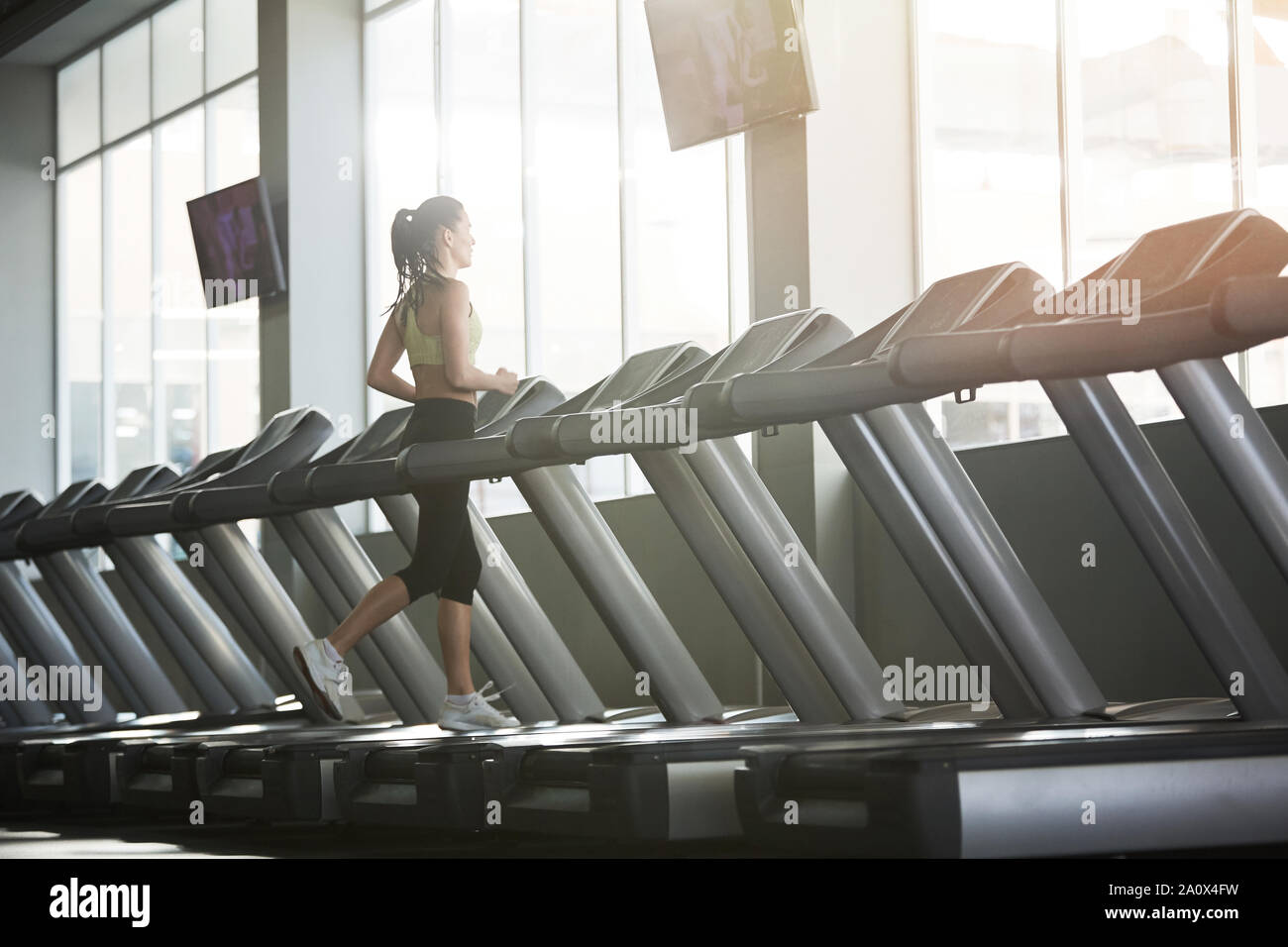 Wide angle back view portrait of young woman running on treadmill alone in empty gym, copy space Stock Photo