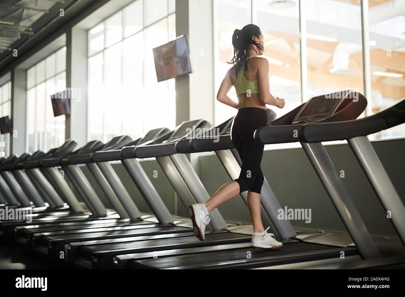 Full length wide angle portrait of young woman running on treadmill alone in empty gym, copy space Stock Photo