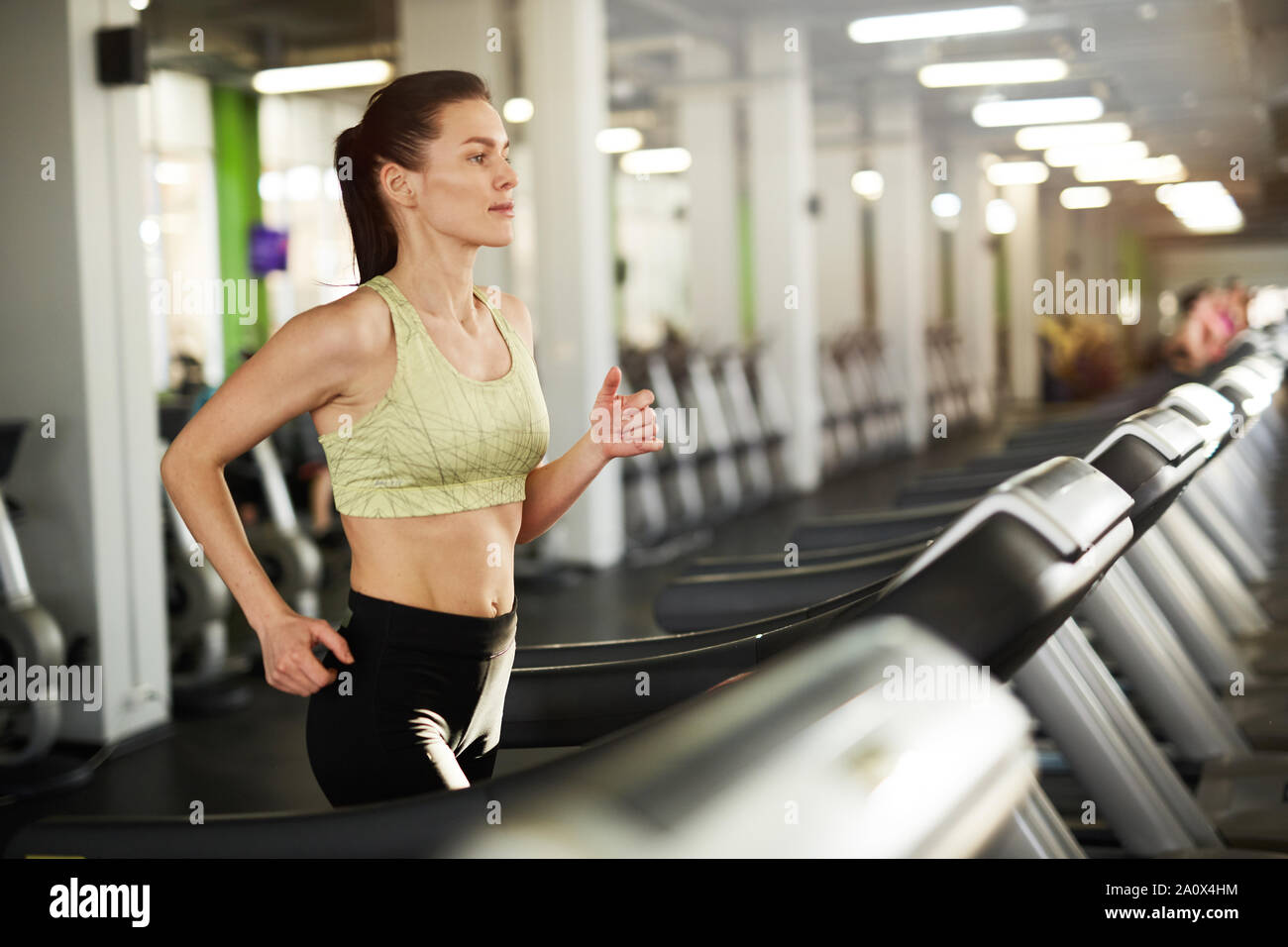 Side view portrait of sweaty young woman running on treadmill during cardio workout in empty gym, copy space Stock Photo