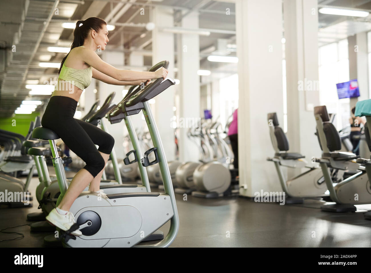 Side view portrait of fit young woman on exercise bike enjoying cardio workout alone in empty gym, copy space Stock Photo