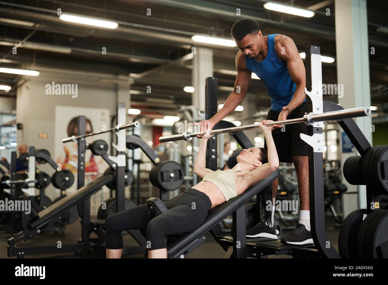 Portrait of African-American fitness coach helping young woman lifting weights on bench in modern gym, copy space Stock Photo