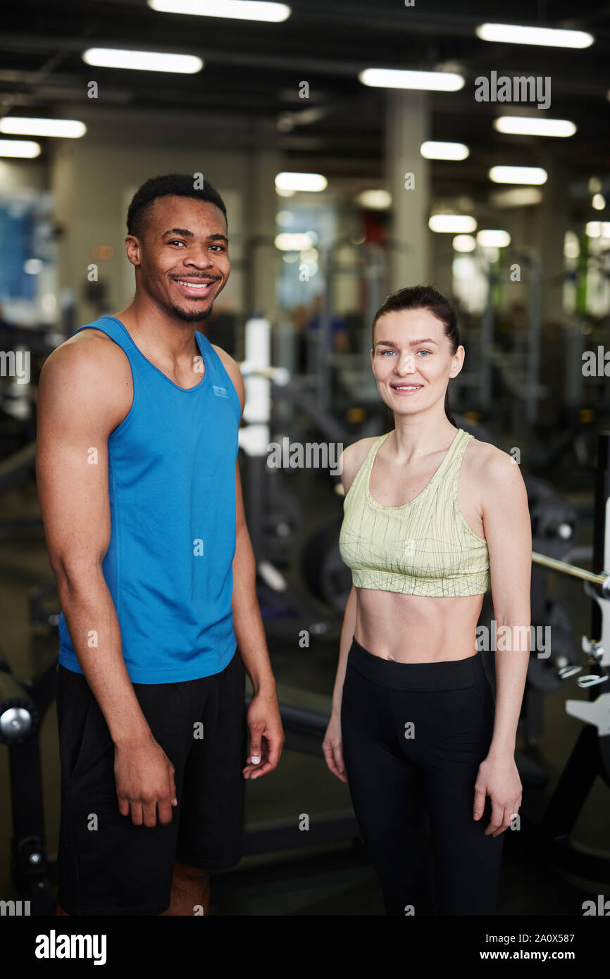 Portrait of fit young couple smiling at camera while posing standing in modern gym, fitness coach concept Stock Photo