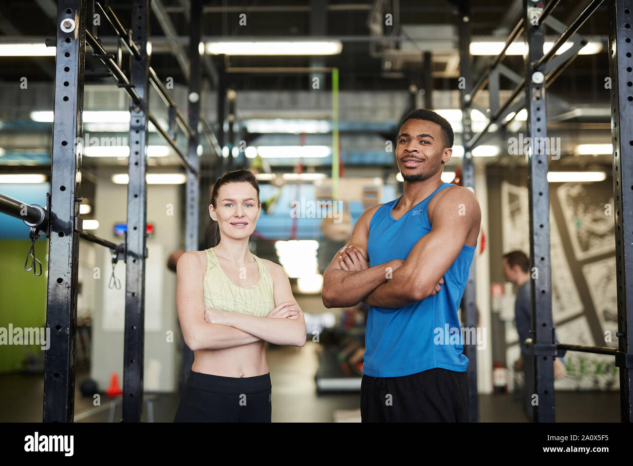Waist up portrait of sportive couple, African man and Caucasian woman, posing confidently standing in modern gym and smiling at camera Stock Photo
