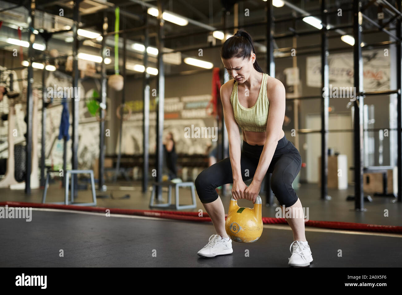 Full length portrait of strong woman lifting kettlebell during cross workout in modern gym, copy space Stock Photo