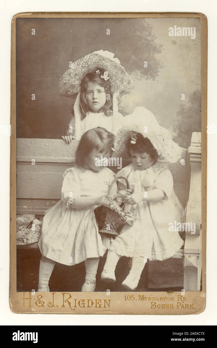 Original antique Edwardian cabinet card, charming studio portrait of three pretty young siblings, sisters, in summer clothes, wearing large straw hats and white smocks, Bowes Park, London, U.K. circa 1904. Stock Photo