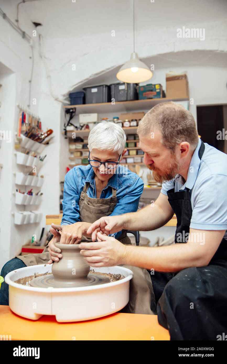Senior woman spinning clay on a wheel with a help of a teacher at