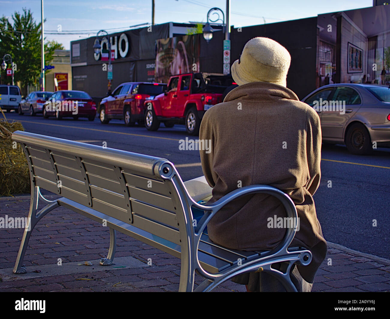 Lady wrapped in warm clothing and a wool hat sits on a bench waiting for the number 85 bus