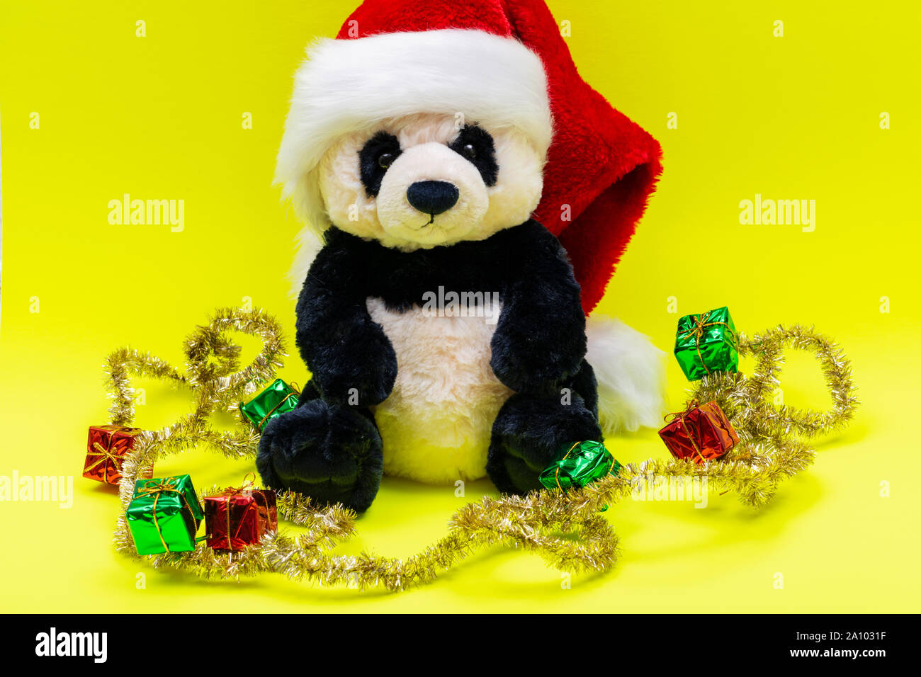 Panda Bear plush toy with Red Christmas Santa Hat surrounded by Tinsel Garlands on bright yellow background. Winter Holidays concept. Stock Photo