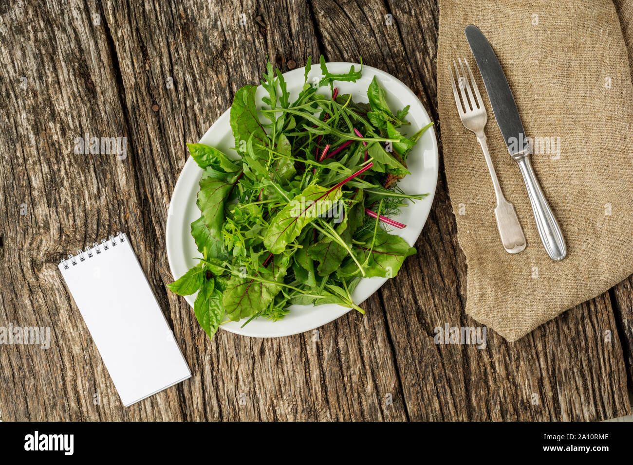 Flat lay. Fresh salad plate with mixed greens on dark wooden background with cutlery and notepad. Healthy food. Green meal. Stock Photo