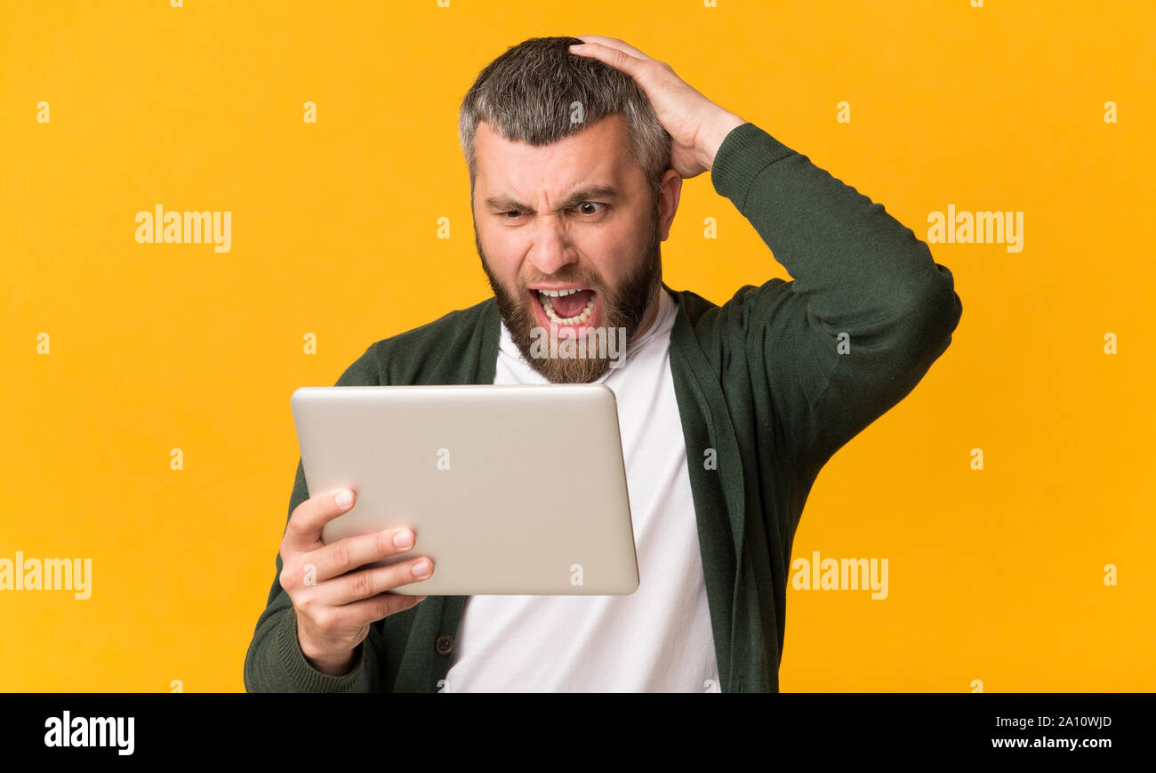 Angry bearded man holding tablet, shouting and touching his head Stock Photo