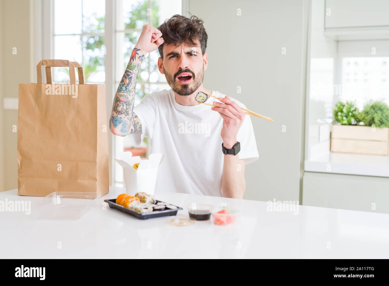 Young man eating asian sushi from home delivery annoyed and frustrated shouting with anger, crazy and yelling with raised hand, anger concept Stock Photo
