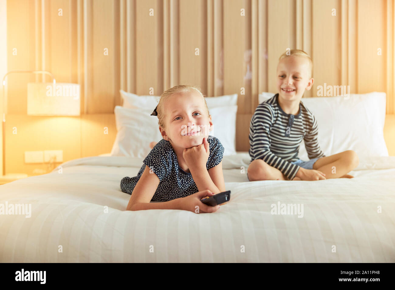 Smiling little girl lying on a bed changing channels while watching televeision with her brother Stock Photo