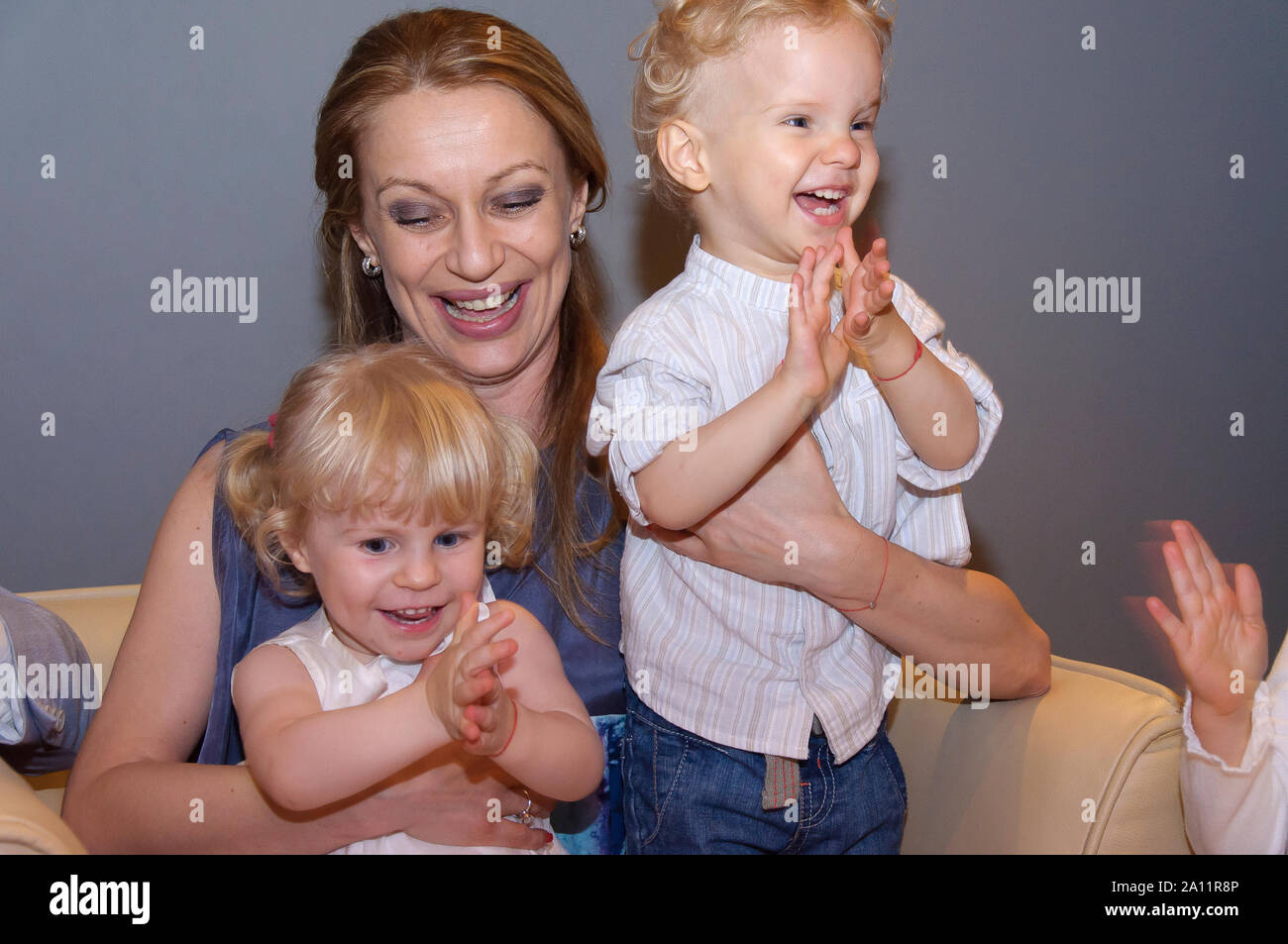 Candid authentic portrait of happy laughing family of mum and two cute little children clapping hands at indoors kid's party Stock Photo