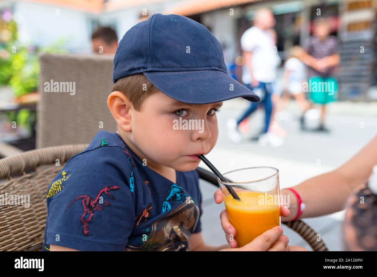 Five-year cute boy in a navy blue baseball cap sitting on a wicker chair outside in a restaurant and drinks orange juice from a drinking straw. Stock Photo