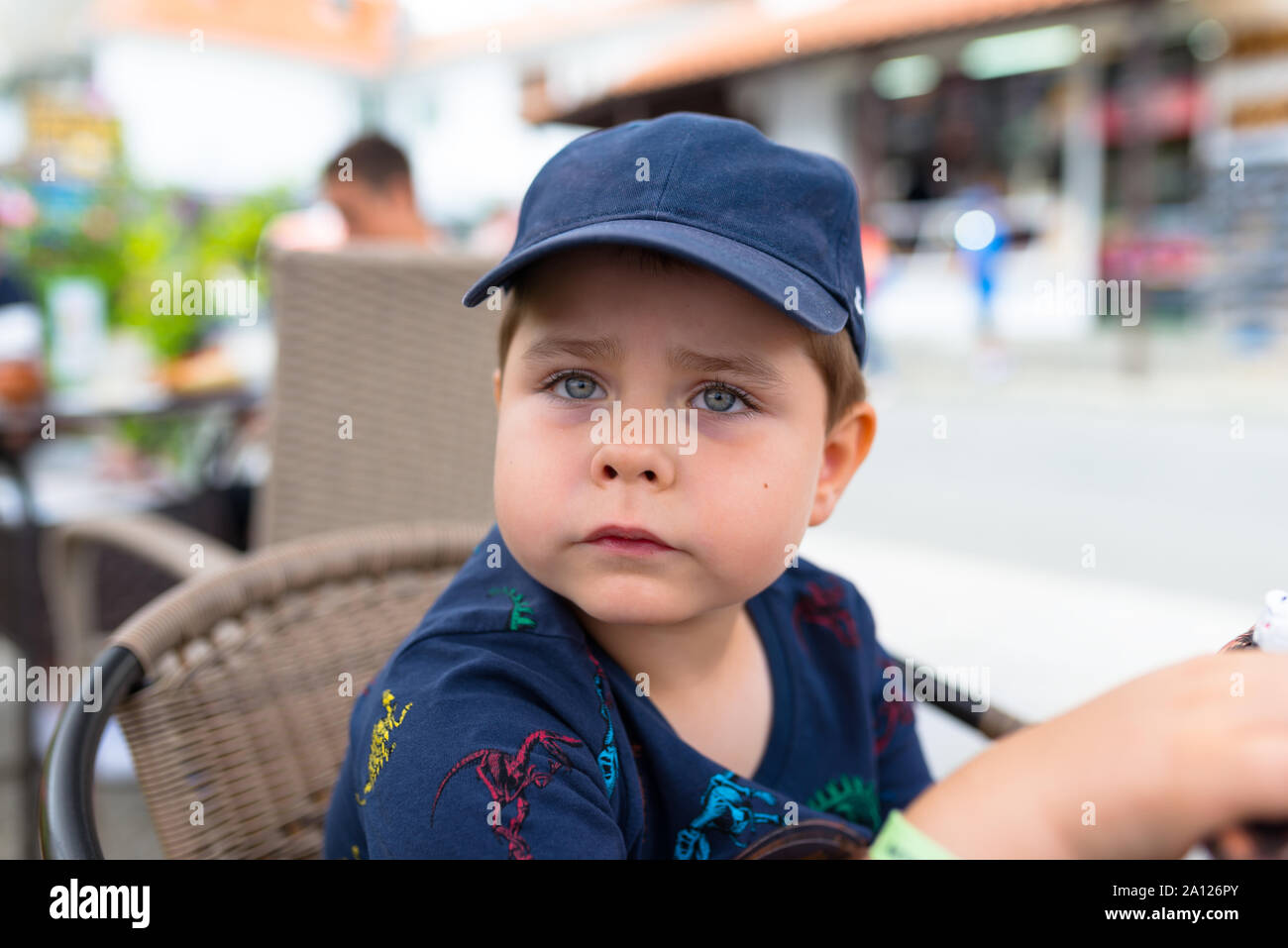 Five-year cute boy in a navy blue baseball cap sitting on a wicker chair outside in a restaurant. Stock Photo