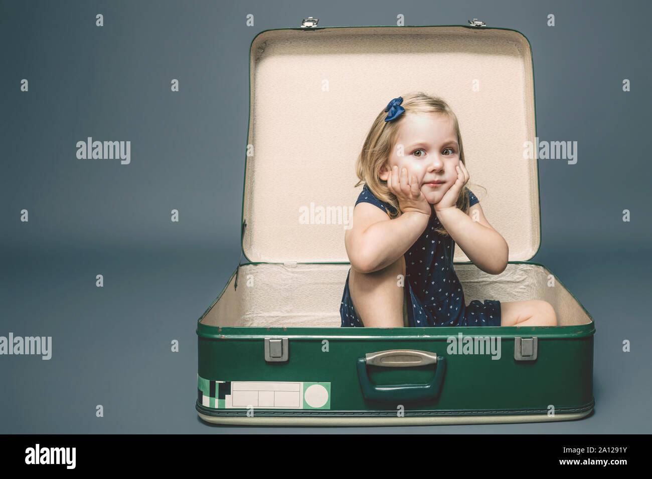 3 year old Caucasian girl with dreamy expression sitting inside a vintage suitcase. Studio shot. Stock Photo