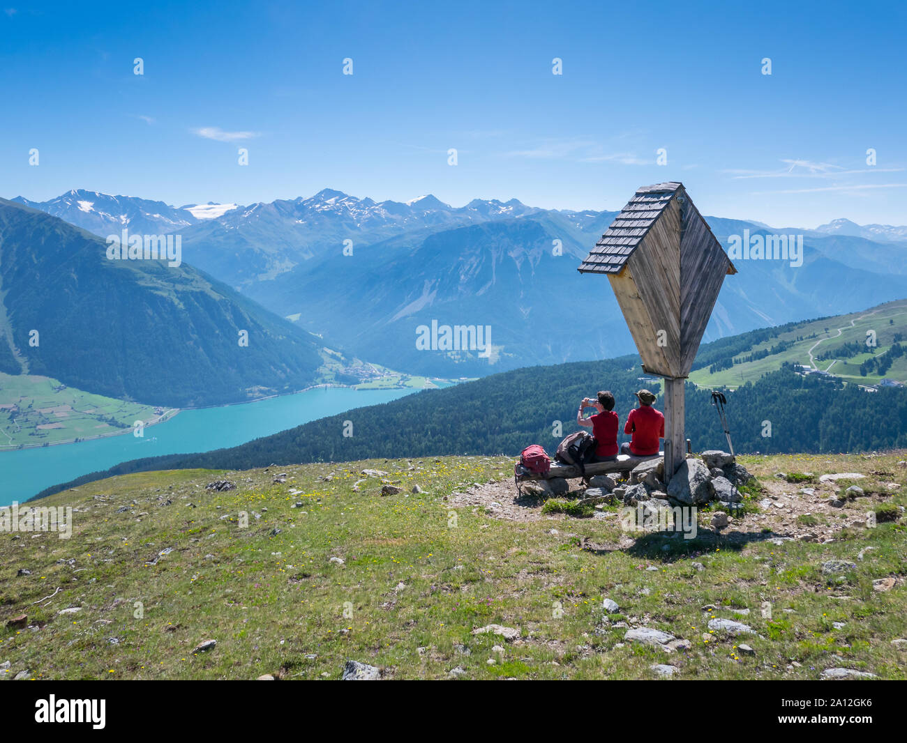 Couple of two medium age hikers resting near a wayside cross above lake Reschen/Resia, Tyrol, Italy Stock Photo