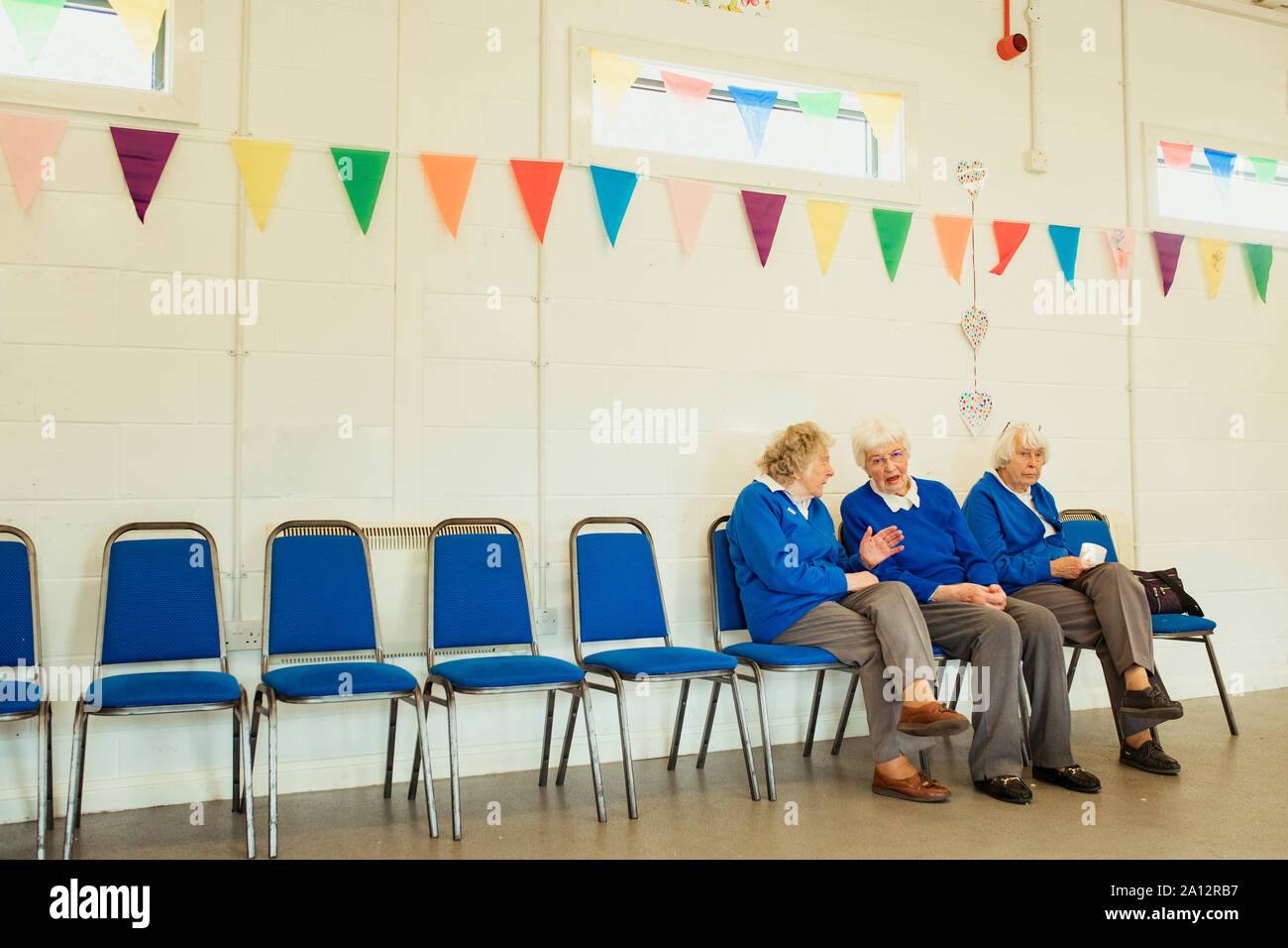 Three senior women talking and sitting indoors on blue chairs in a hall decorated with bunting. Stock Photo