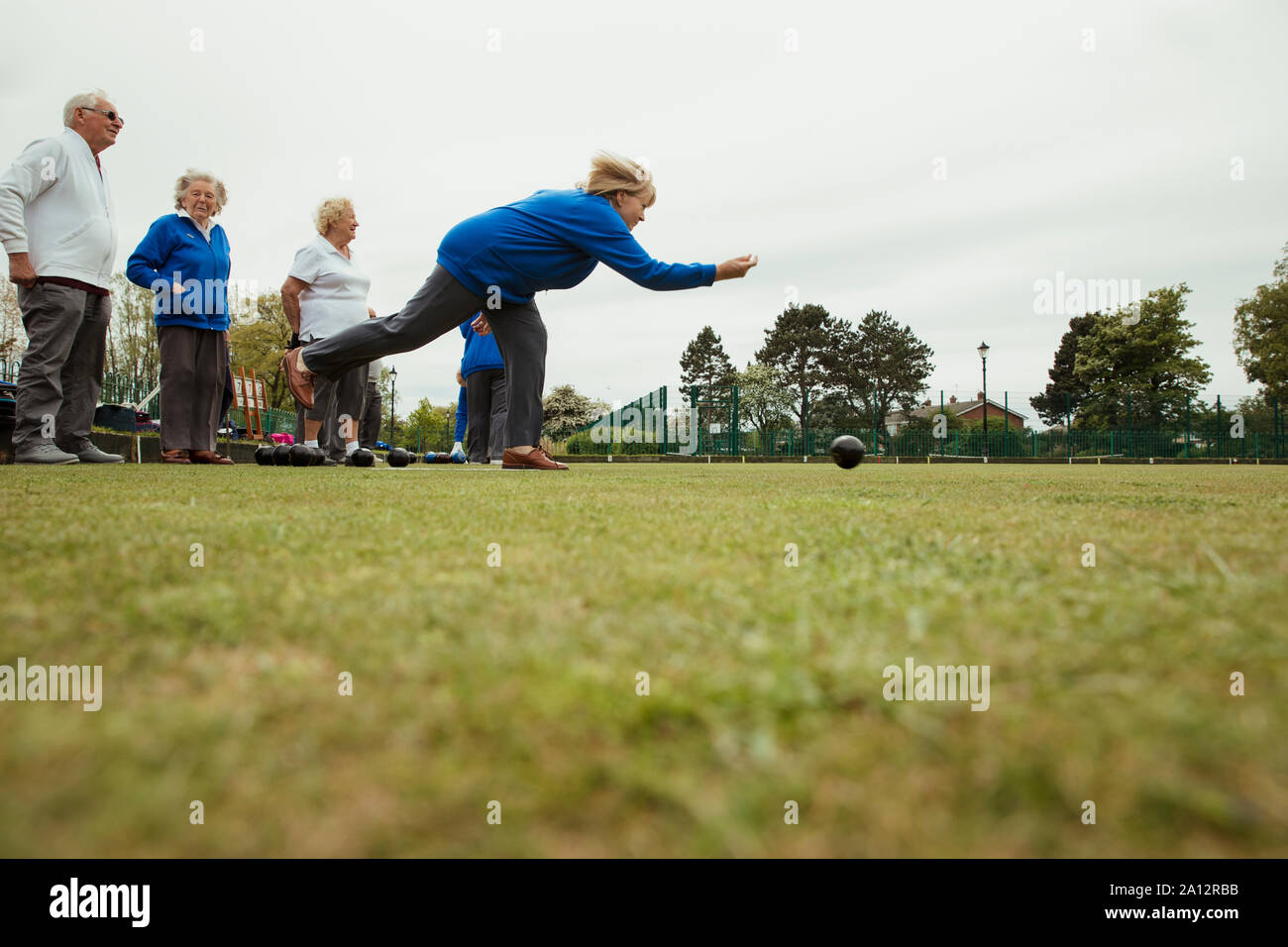 A side view shot of a group of senior friends lawn bowling on grass, with one woman who has taken her shot. Stock Photo