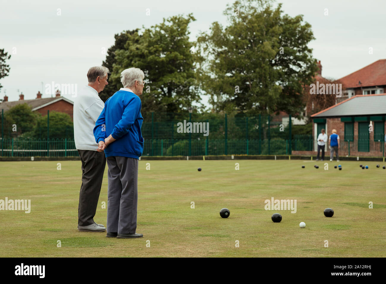 A rear view shot of senior adults spectating a lawn bowling game, standing with their hands behind their back. Stock Photo
