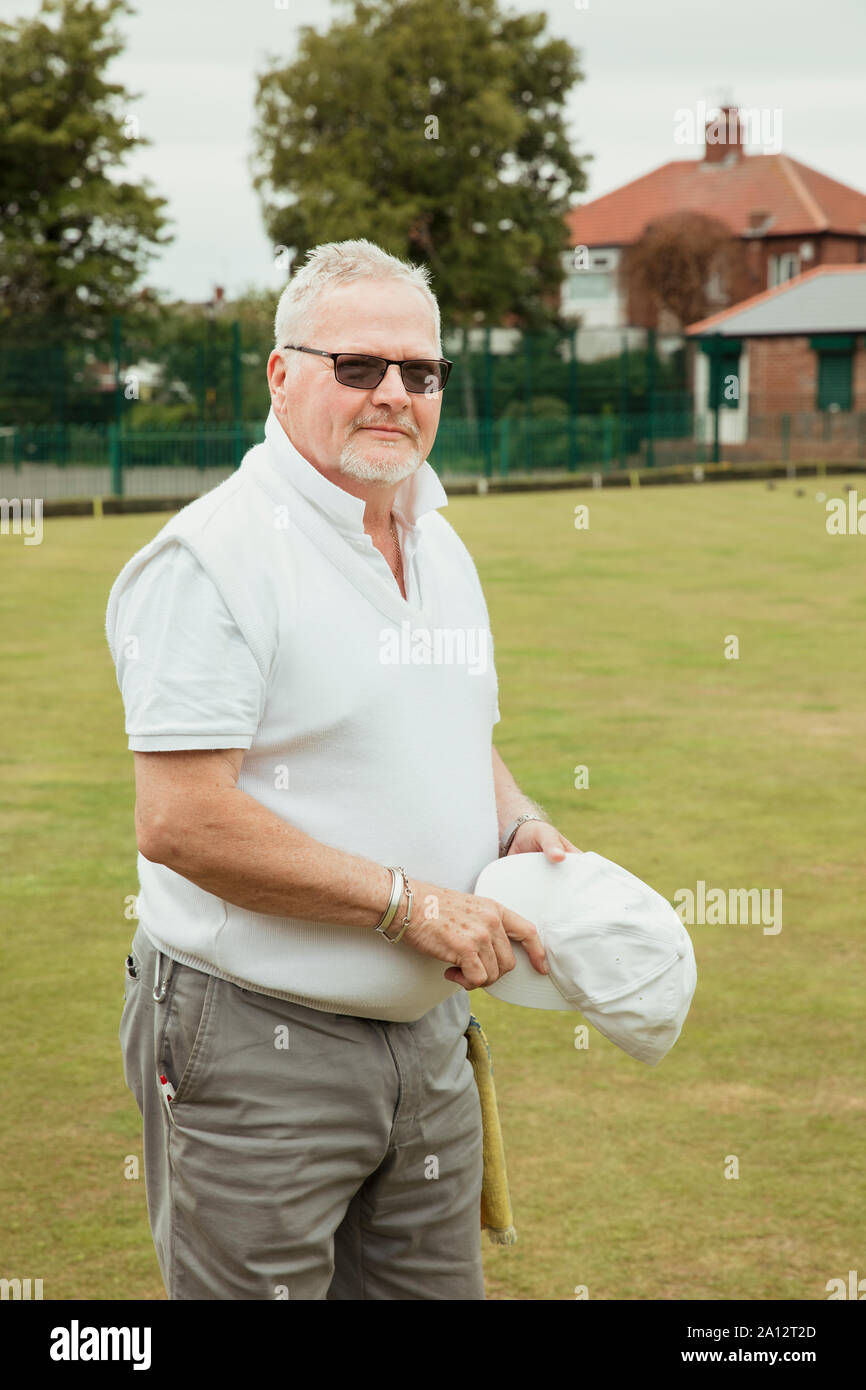 A portrait of a senior man smirking at a bowling green. He is wearing sunglasses and is holding a white cap. Stock Photo