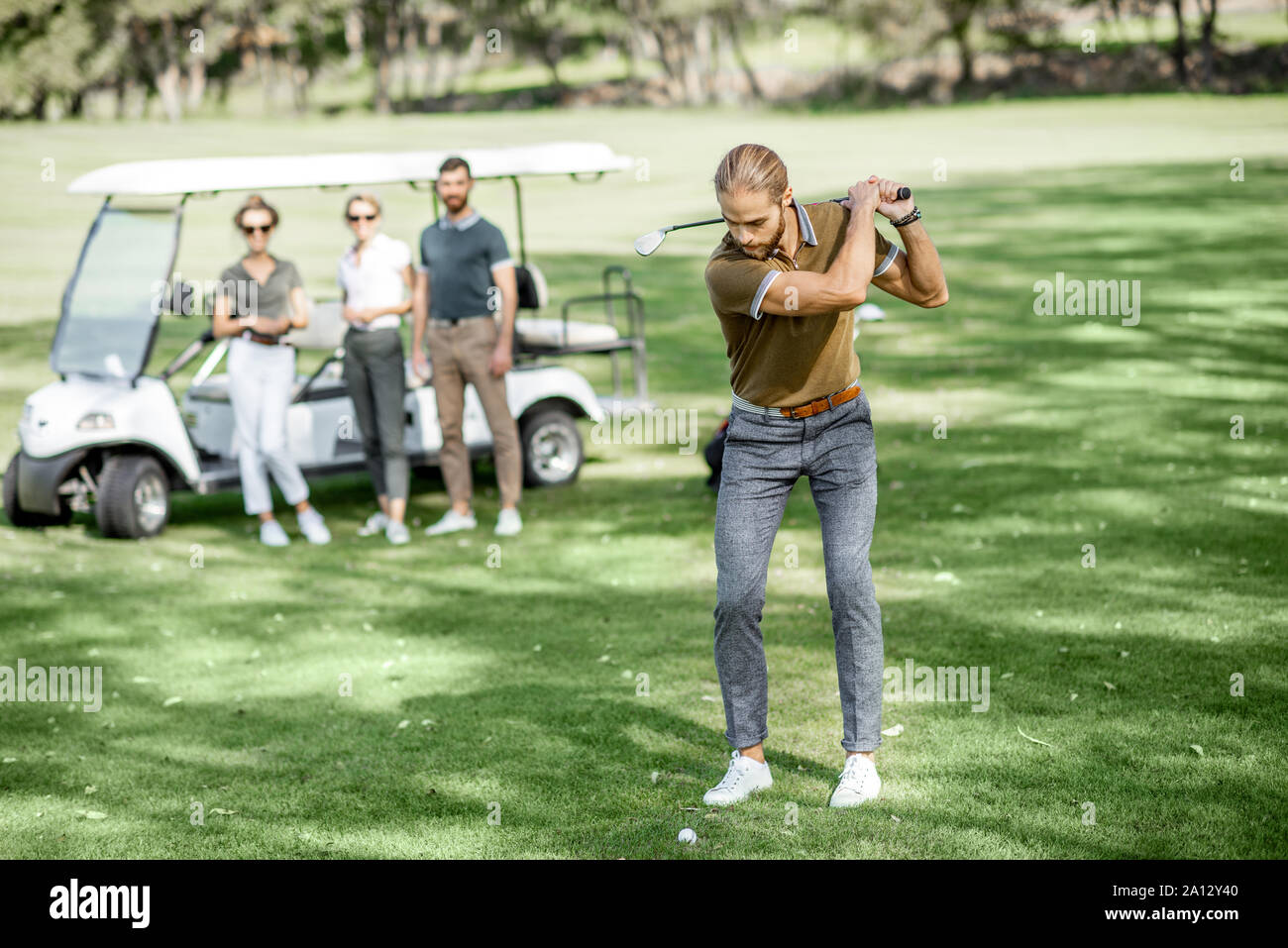 Handsome man swinging with putter, playing golf on the course with friends and golf car on the background on a sunny weather Stock Photo