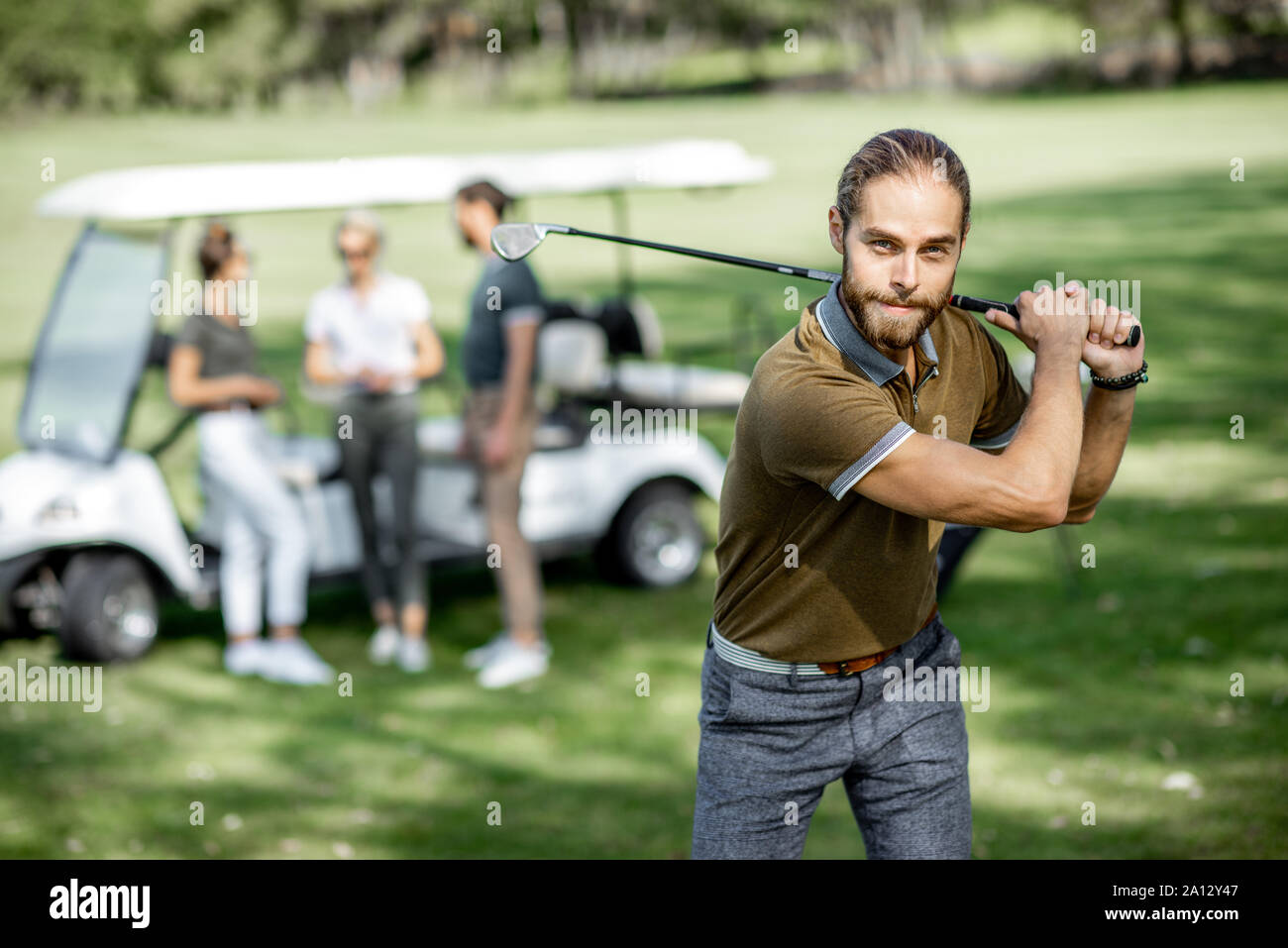 Portrait of a handsome man swinging with putter, playing golf on the course with friends and golf car on the background on a sunny weather Stock Photo