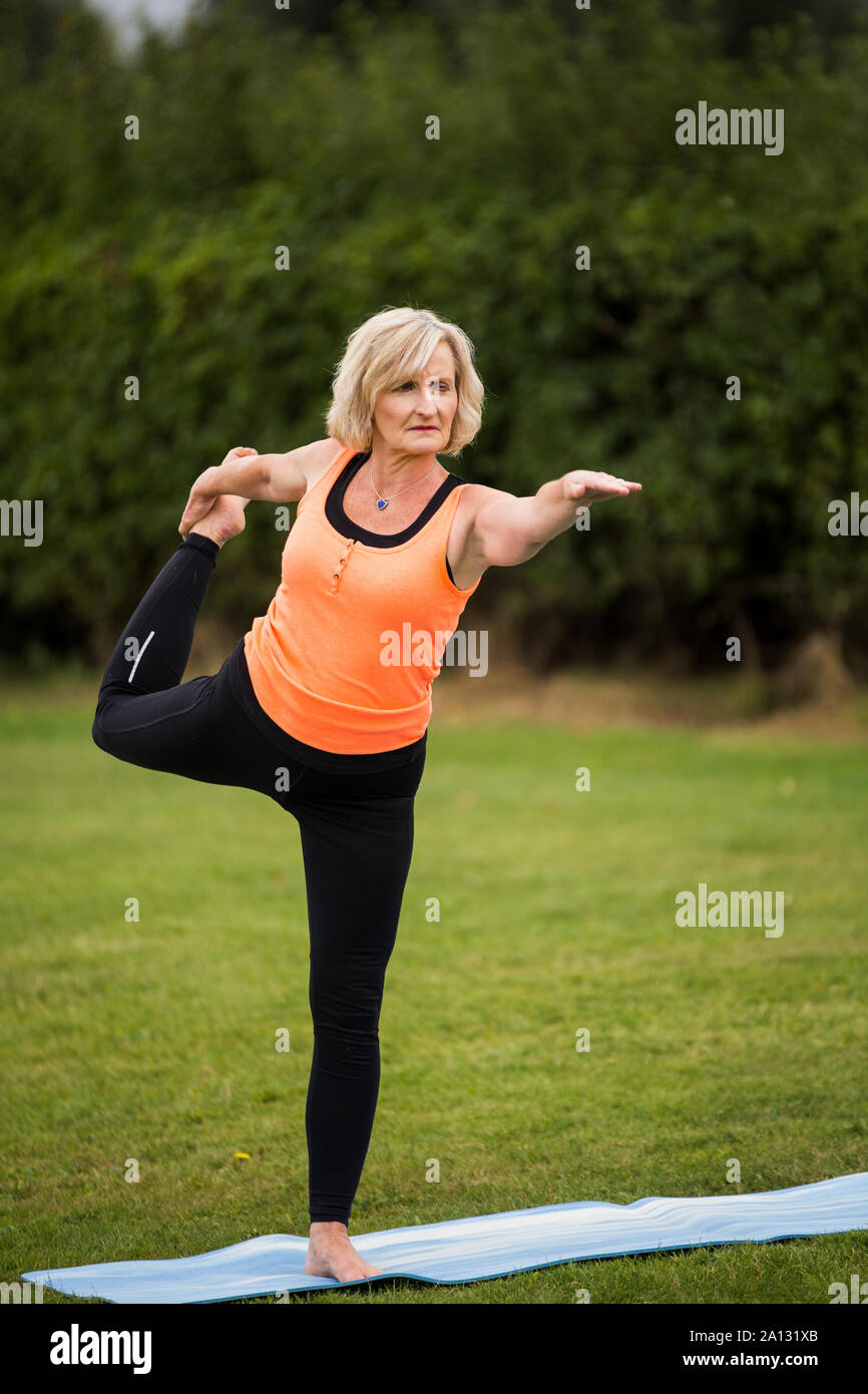 A middle aged woman practicing yoga barefoot outside in a grassy