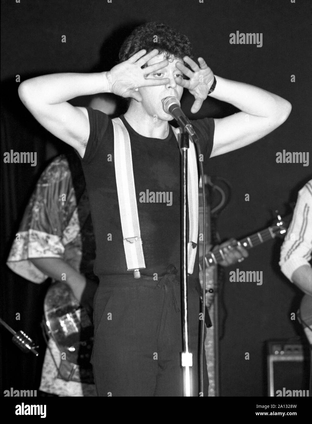 Lou Reed performs onstage at the Bottom Line in March, 1978. He is peeking through his fingers while standing at the microphone in suspenders. Stock Photo