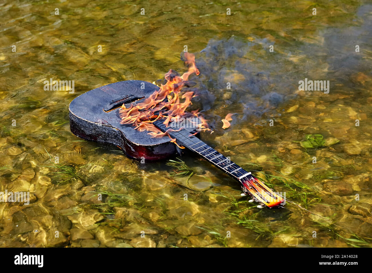 An ignited guitar floats on riverbank, fire burns on its surface. From the flame comes black smoke. The strings are torn. A musical instrument drifts Stock Photo