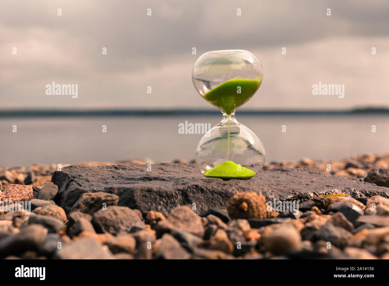 Hourglass with green sand on the lake, stand on small stones. Time concept Stock Photo