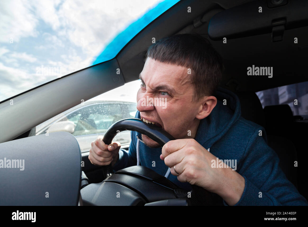 An unbalanced, goosey man bites a car steering wheel from anger while