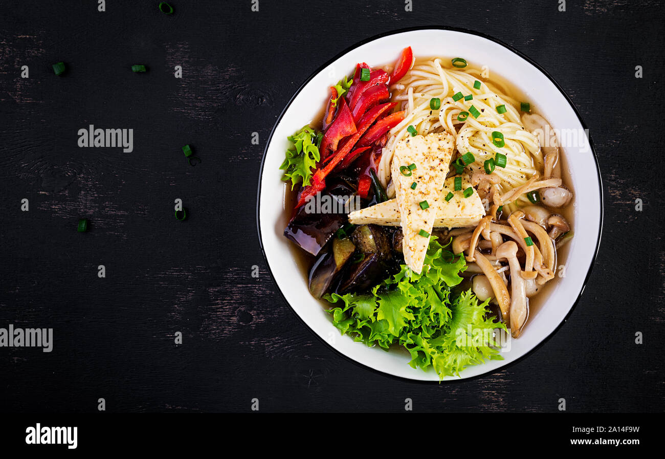 Asian vegan noodles soup with tofu cheese, shimeji mushrooms and fried eggplant in bowl on dark background. Top view Stock Photo
