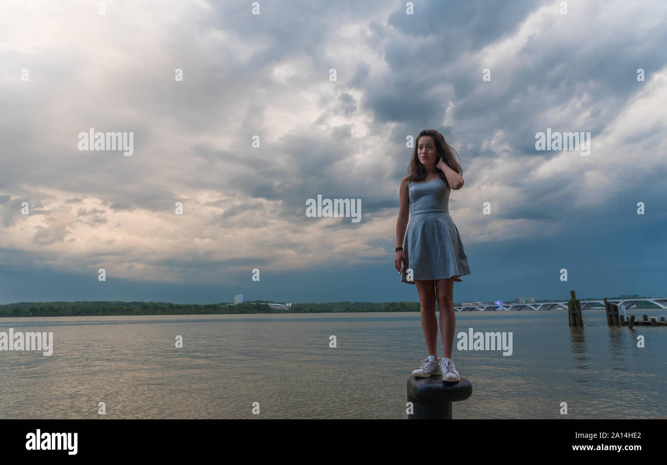 Teen girl in dress standing on bollard in front of Potomac River with mobile phone in her hand Stock Photo