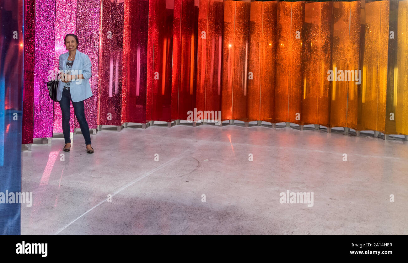 Asian woman in blue jacket and black pants standing in front of colorful mirrors Stock Photo