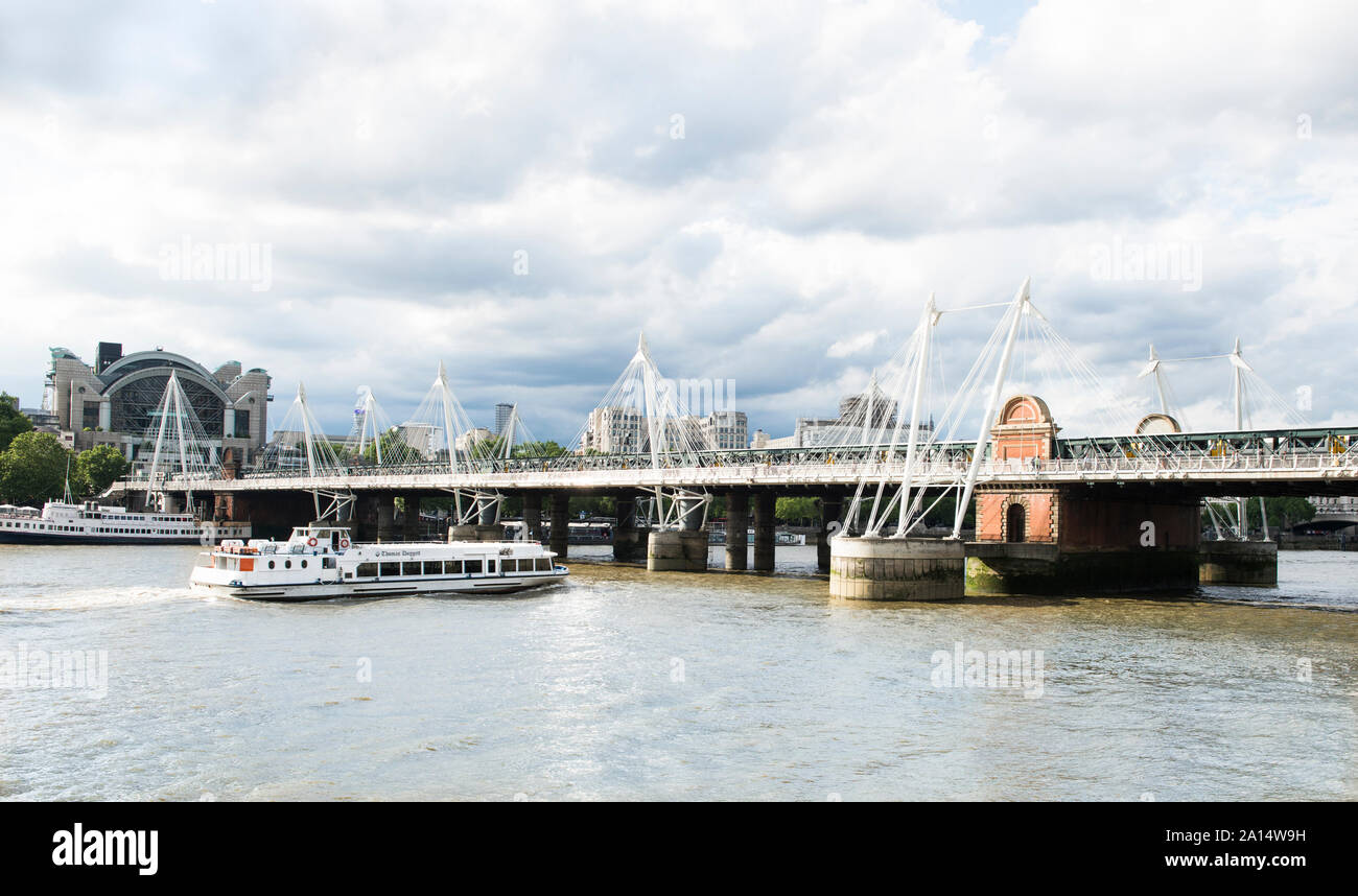 Golden Jubilee Bridges and Charing Cross Station. A touring boat is about to pass under the bridge. Hungerford Footbridges. Stock Photo