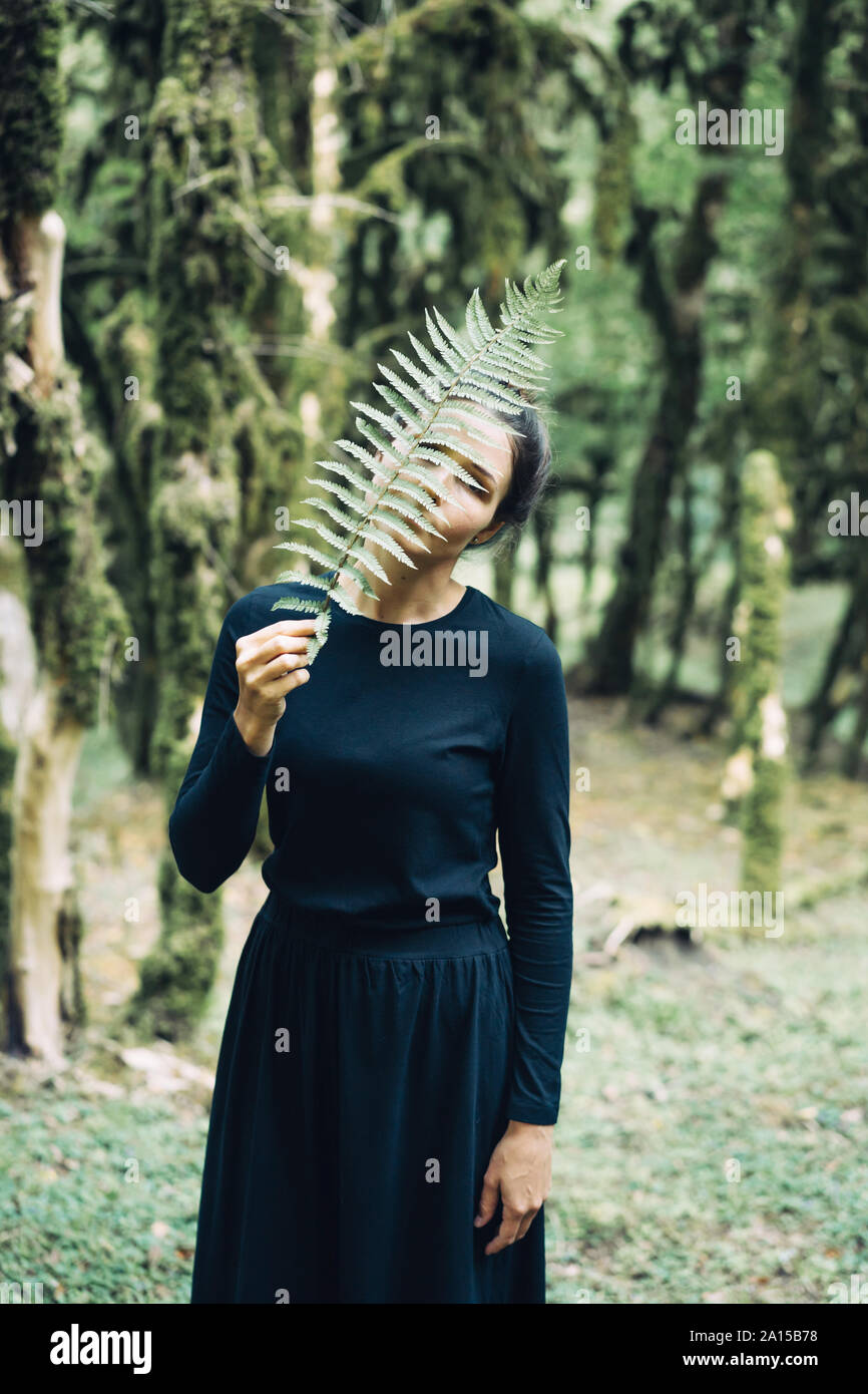 A mysterious woman in a long black dress in the forest covers her face with a fern branch. Stock Photo