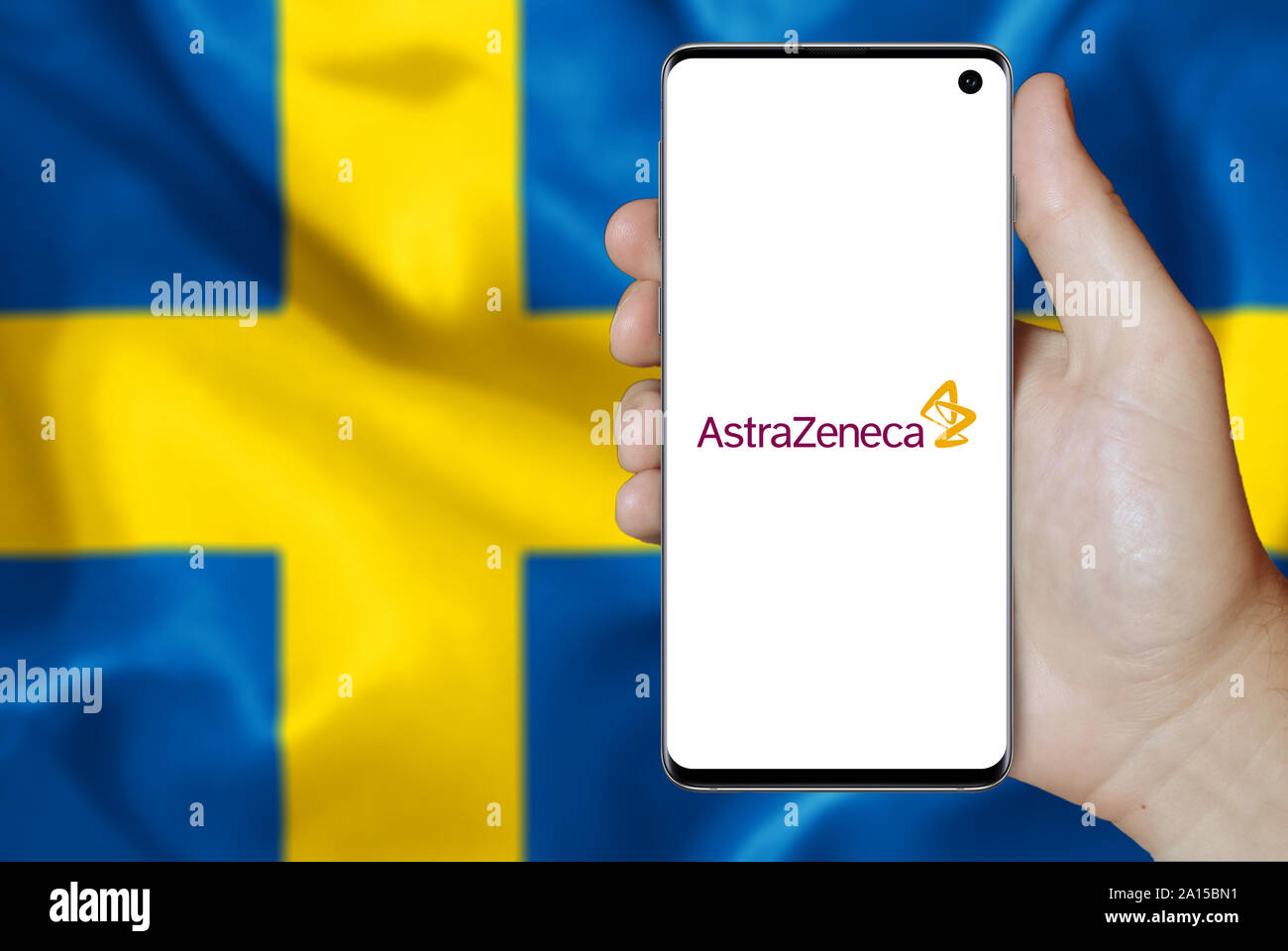 A man holds a smartphone displaying the logo of company AstraZeneca listed on OMX Stockholm. Swedish flag in the background. Credit: PIXDUCE Stock Photo
